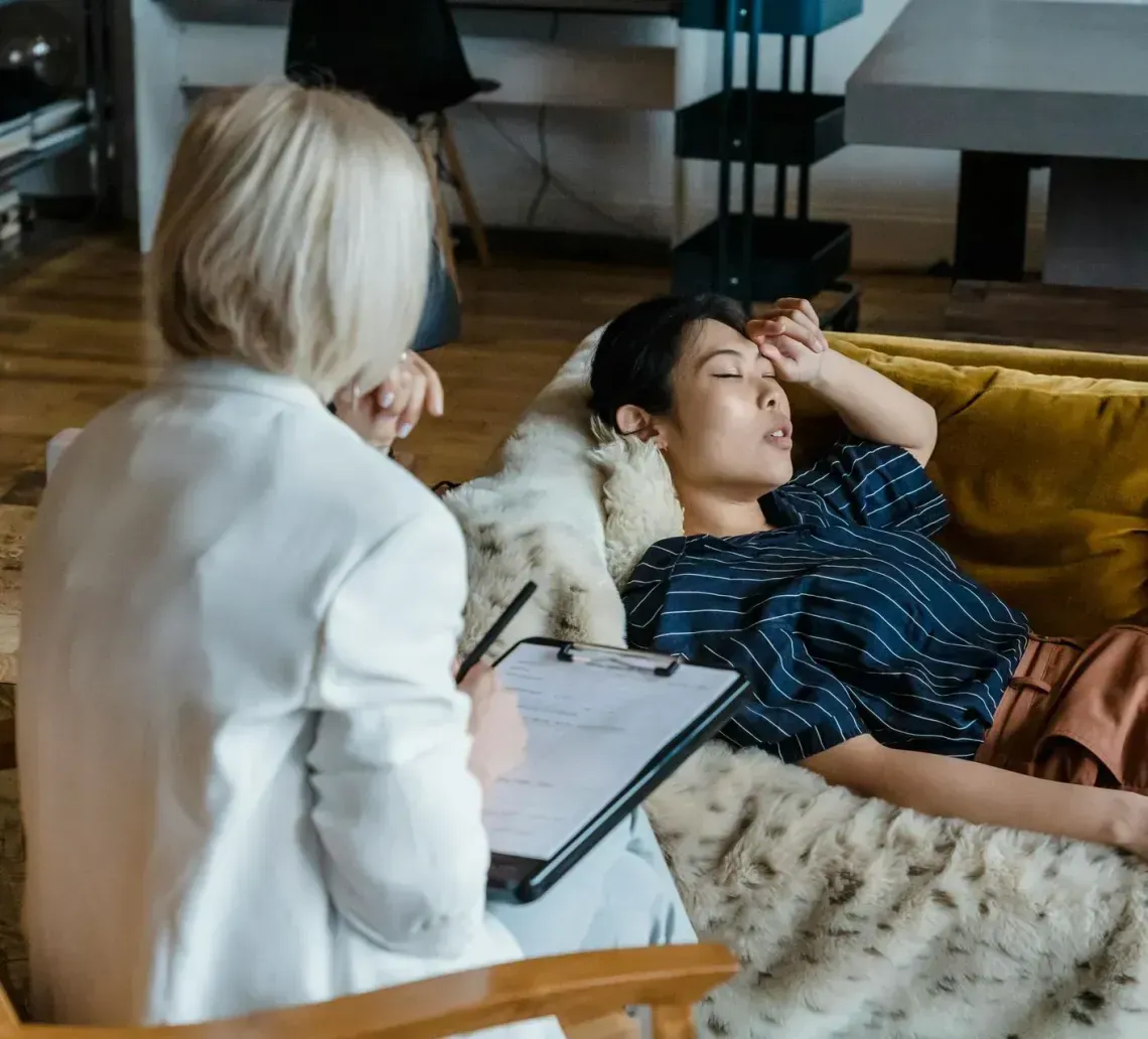 A therapist in a white jacket takes notes on a clipboard while a person lies on a sofa with their hand to their forehead.