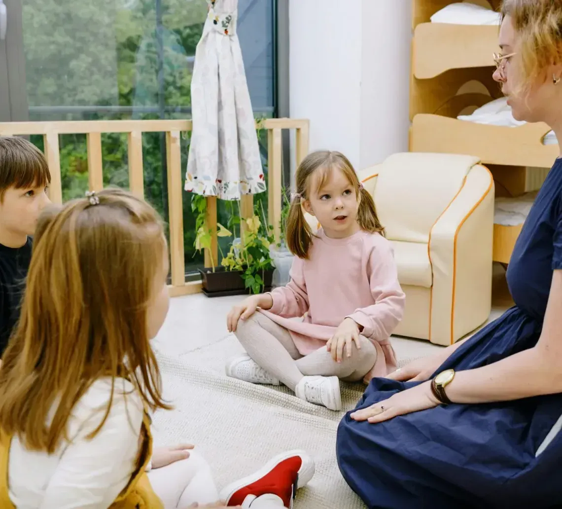 A teacher sits on the floor in a classroom, talking to three children who are gathered around in a circle.