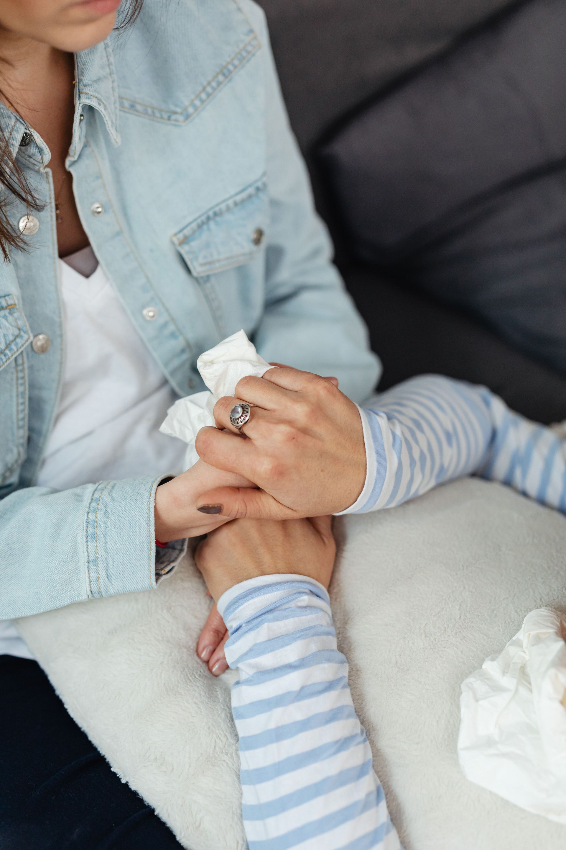 Adult holding a baby’s hand with a tissue, wearing a light denim shirt on a couch