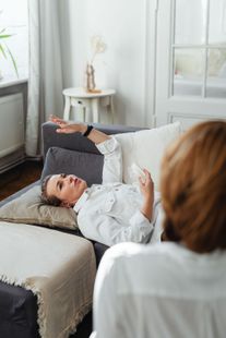 Person reclining on a couch in a bright living room, speaking with someone seated nearby.