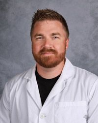 A man with a beard is wearing a white lab coat and a black shirt.