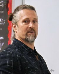 A man with hair pulled back and a beard is standing in front of a wall.