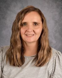 Portrait of a smiling woman with wavy brown hair against a gray studio backdrop, wearing a striped shirt