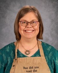 Smiling woman wearing glasses and apron reading “But did you read the” against a gray backdrop