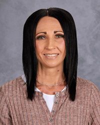 A woman with long brown hair is wearing a brown sweater and white shirt and smiling for the camera.