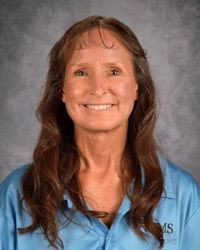 A woman with long brown hair is wearing a lighter blue shirt and smiling for the camera.