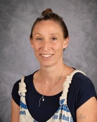 Smiling person in navy shirt and white overalls against a gray studio backdrop