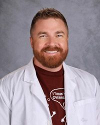 A man with a beard is wearing a white lab coat and a maroon shirt.