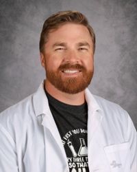 Smiling man in a white lab coat and black T-shirt against a gray studio backdrop