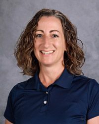 A woman with curly hair is wearing a dark blue polo shirt anad smiling for hte camera
