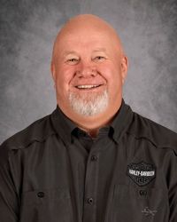 Smiling bald man in a dark button-up shirt with a logo, against a gray studio backdrop