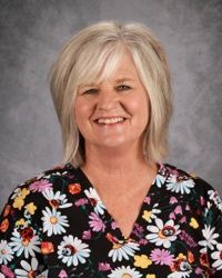 Smiling person with blonde hair in a black floral blouse against a gray studio backdrop