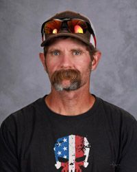 A man with a mustache is wearing a hat and sunglasses, black shirt with red,white and blue.
