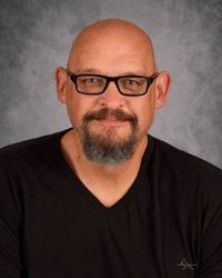 Portrait of a bald man with glasses and a goatee, wearing a black shirt against a gray backdrop