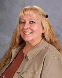 A woman with long blonde hair is wearing a tan shirt and smiling for the camera.