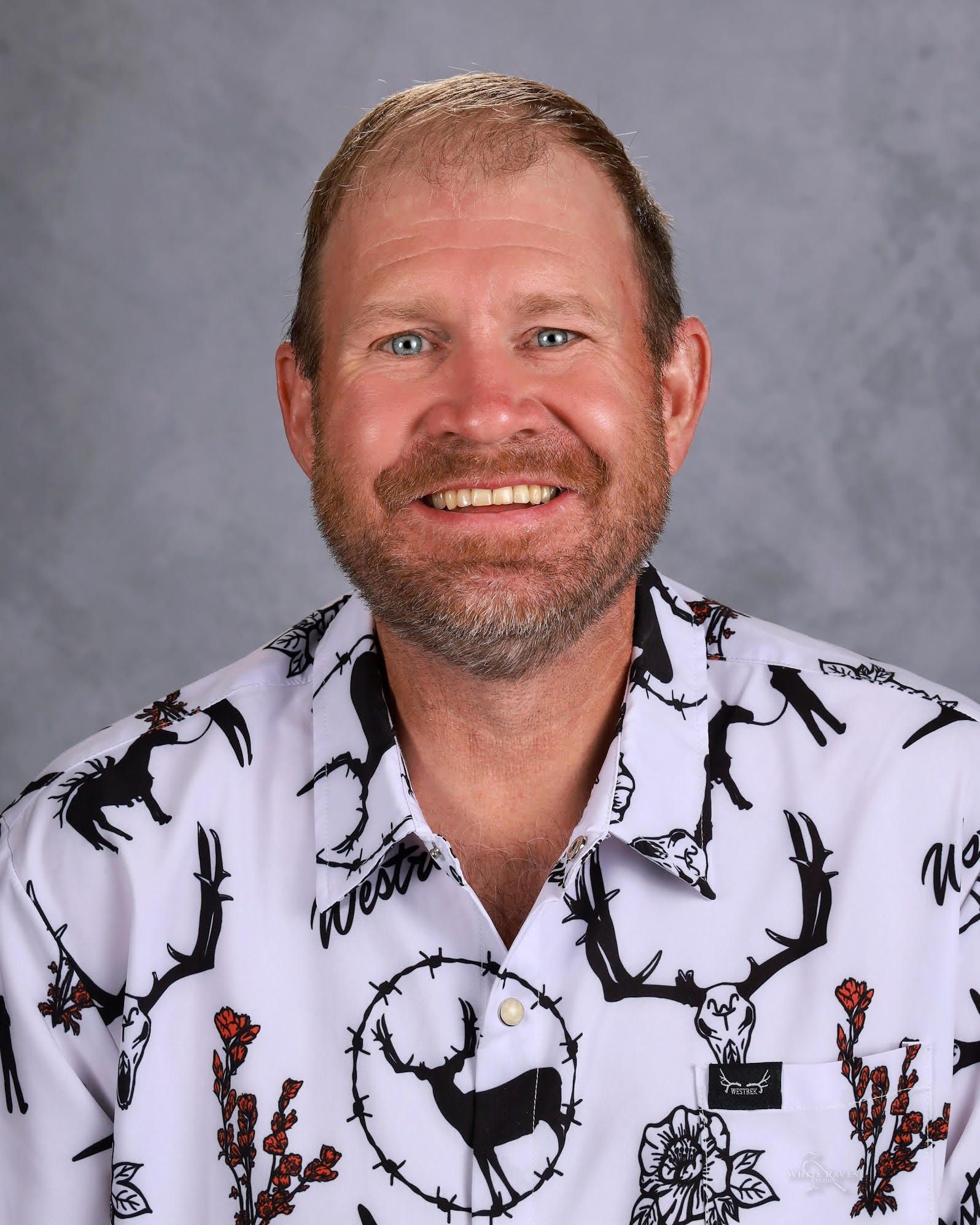 Man with a beard smiling, wearing a patterned white shirt with black deer and red floral designs.