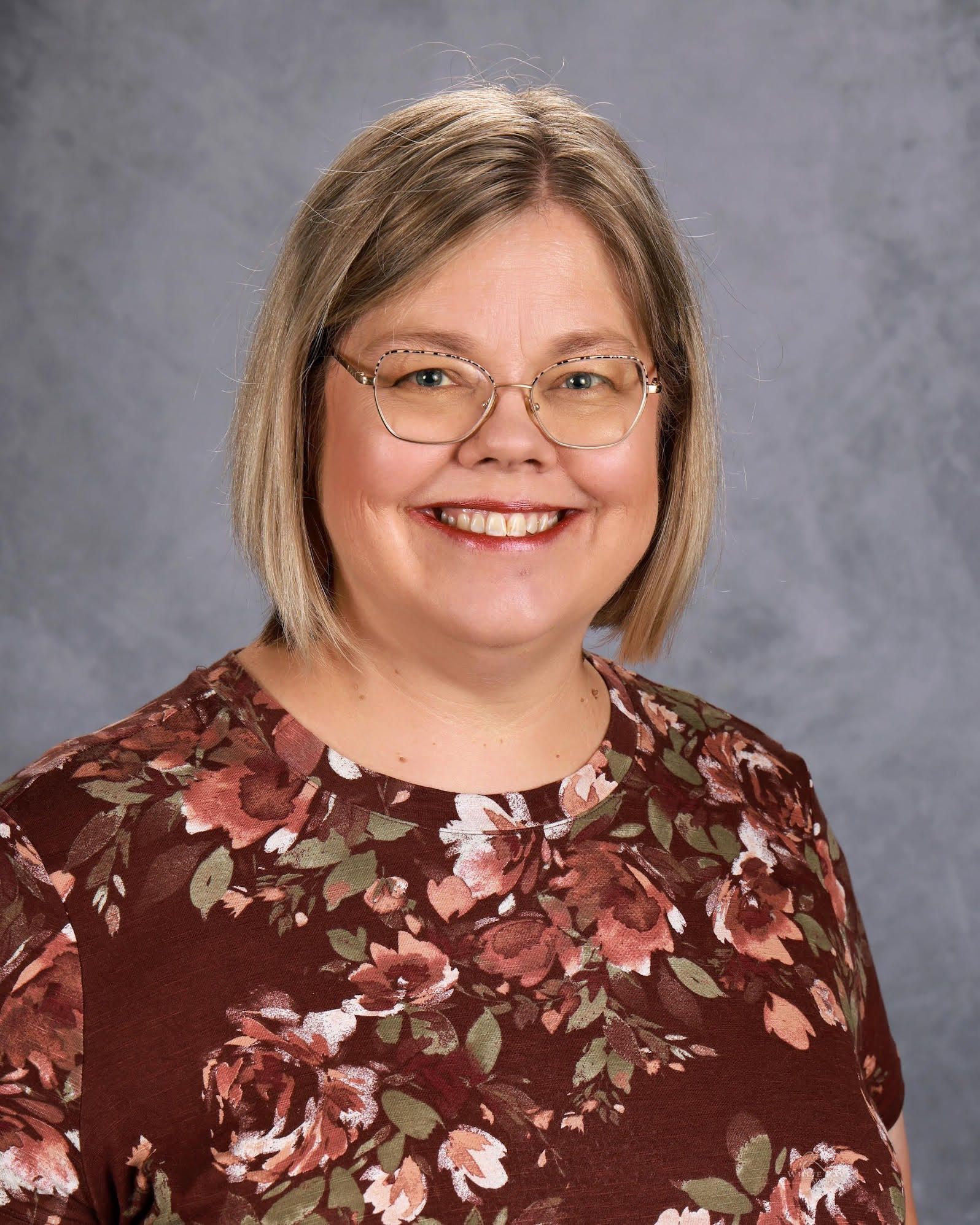 Woman with blonde hair and glasses smiling, wearing a floral shirt.