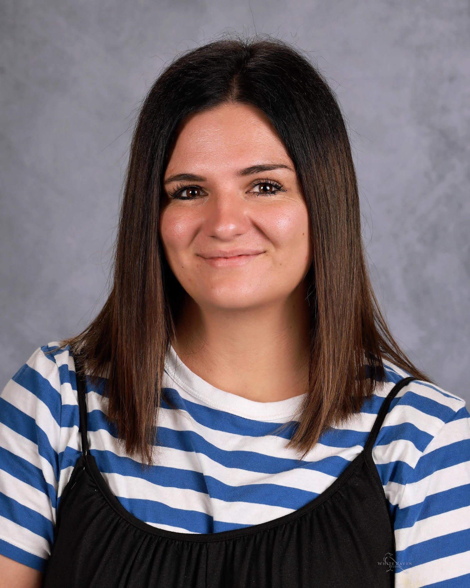 Woman with brown hair, smiling, wearing a blue and white striped shirt under a black top, gray background.