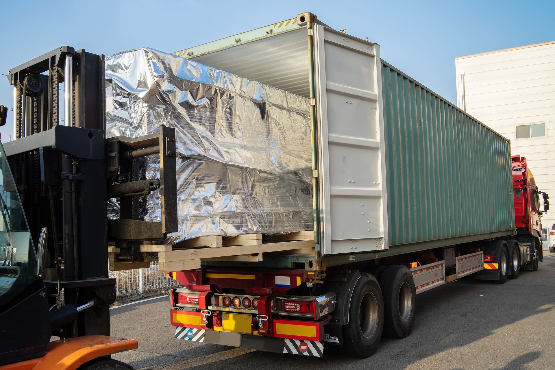 Carrello elevatore che carica un pallet di merci avvolto in un involucro argentato in un container verde su un semirimorchio.