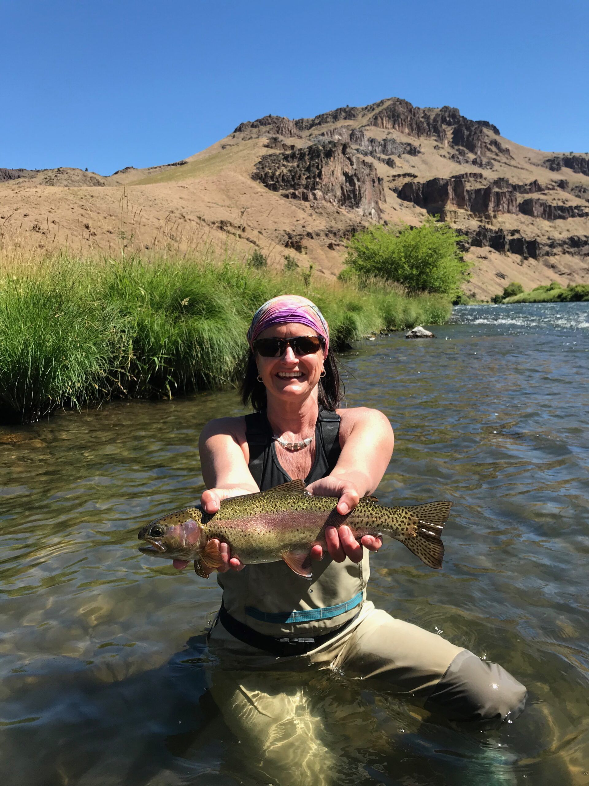 A woman is kneeling in the water holding a rainbow trout.