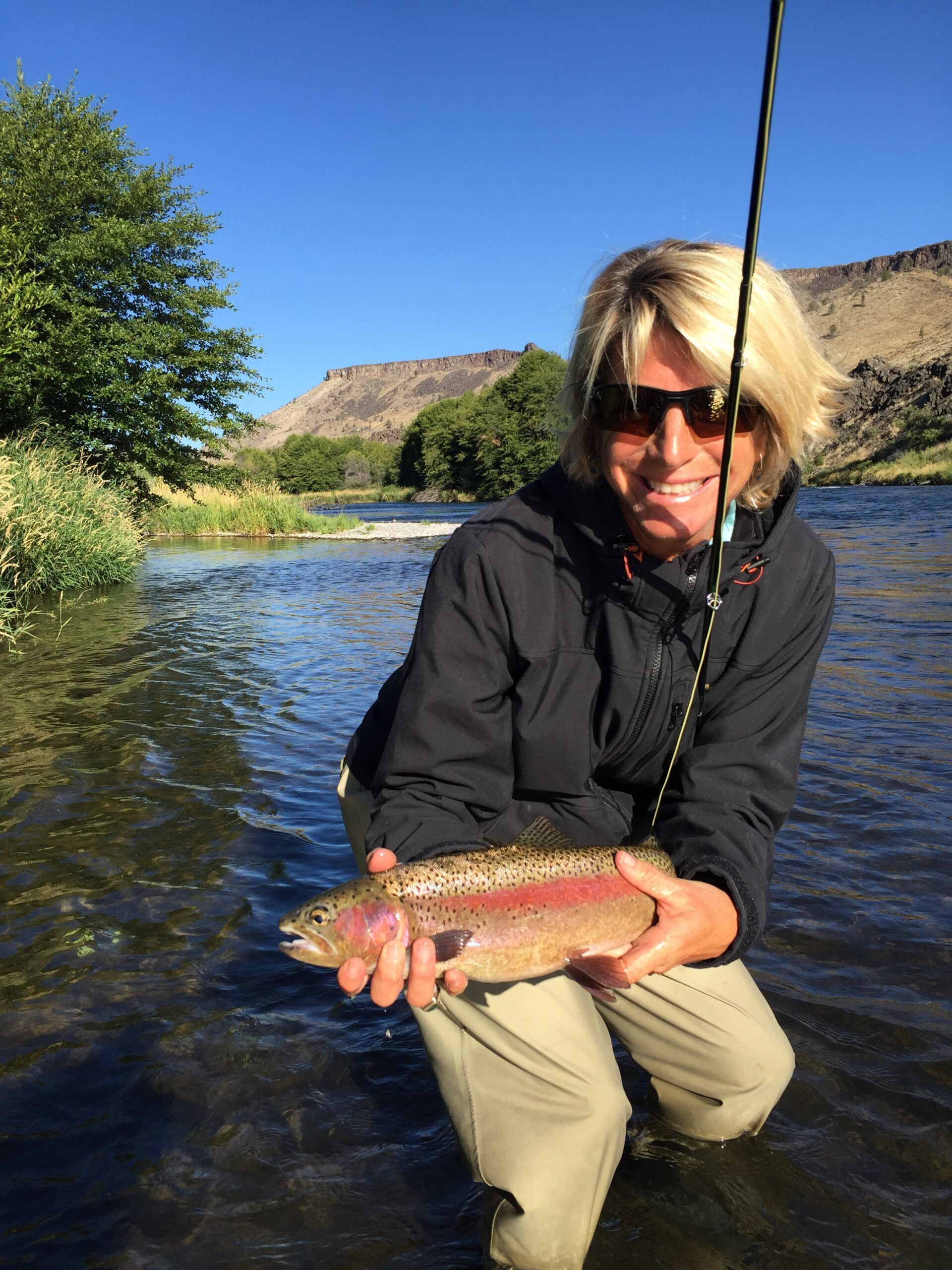 A woman is kneeling in the water holding a rainbow trout.