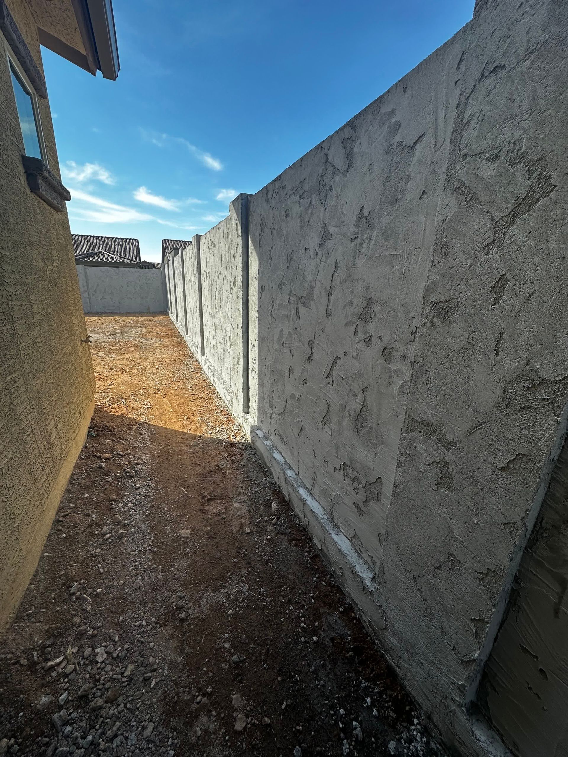 A concrete wall surrounds a dirt path between two buildings.