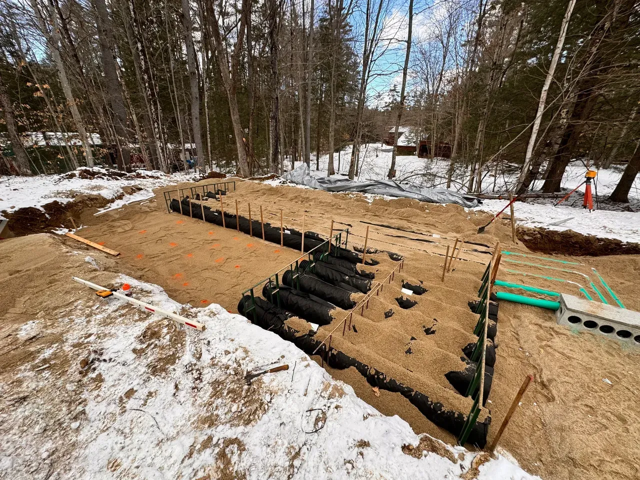 Construction site with trench and sand-filled foundation for a structure, surrounded by snow and trees.