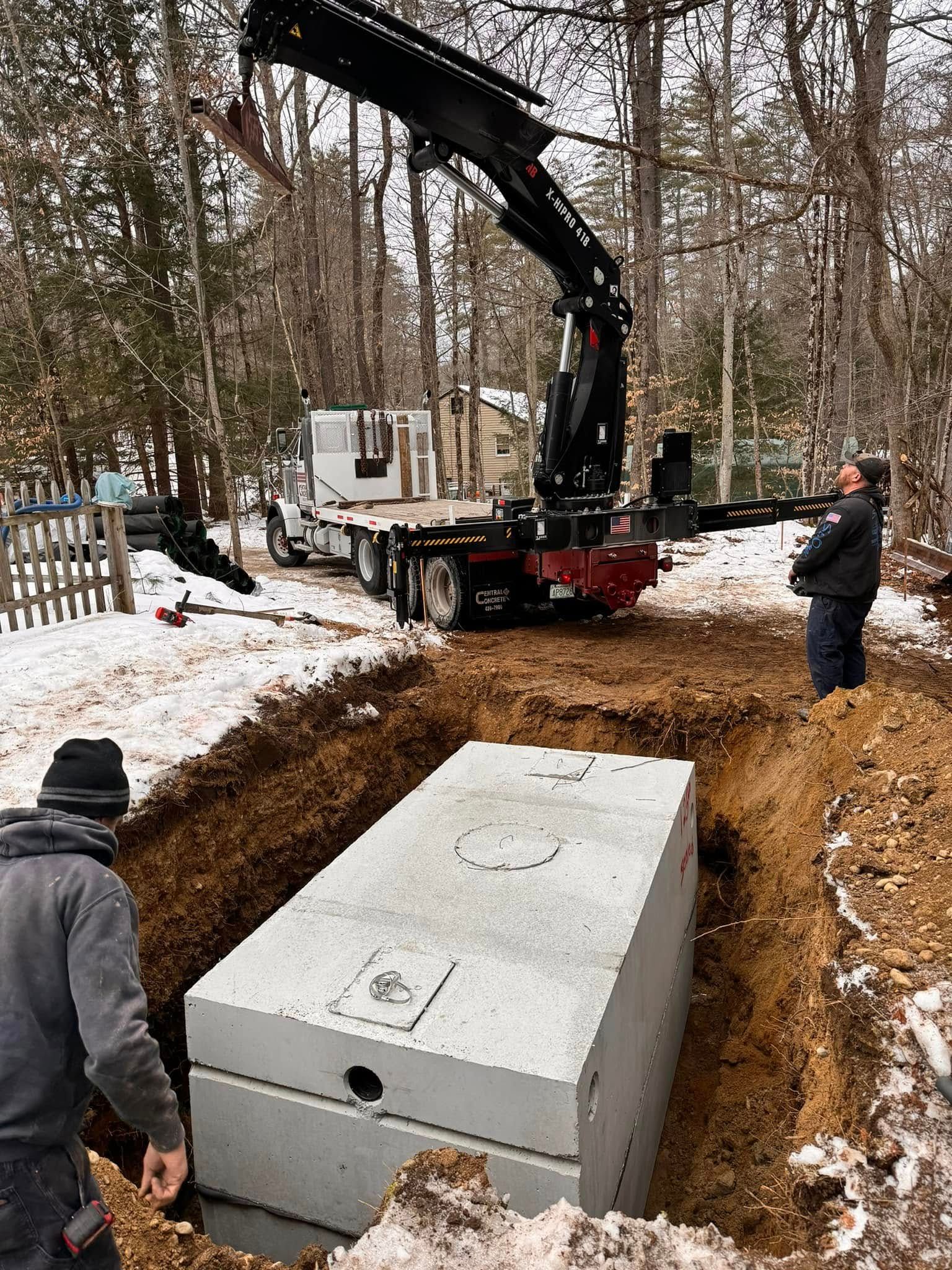 Crane lowering a concrete septic tank into a prepared trench in a snowy wooded area. Two workers observe.