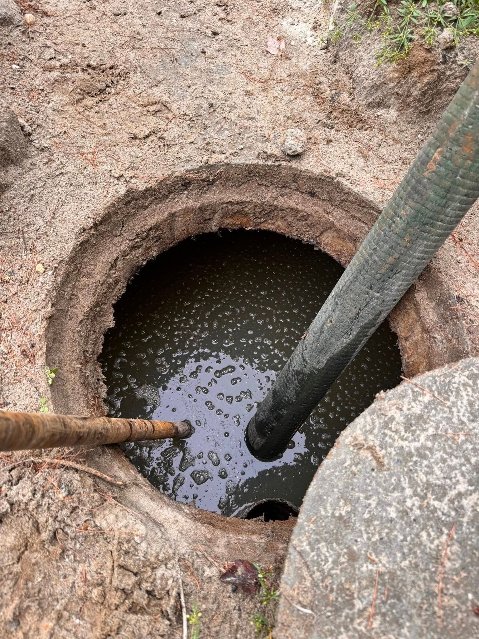 Sewer hole with hoses; dark water inside. Outside, dirt and rock.