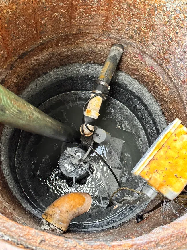 Inside a circular, concrete pit, a sump pump sits in murky water with attached pipes and electrical components.