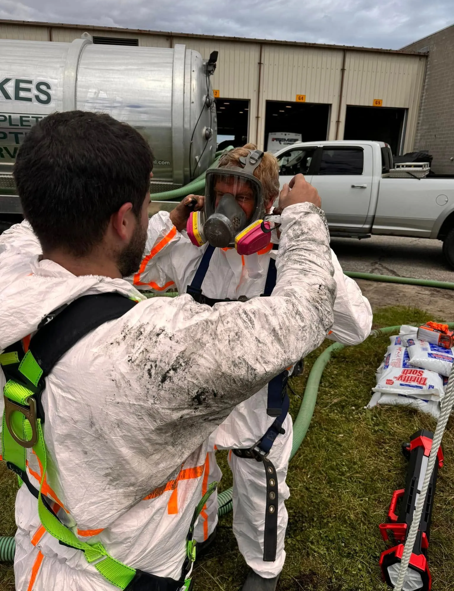 Two workers in safety gear fitting a respirator mask outdoors near trucks and equipment