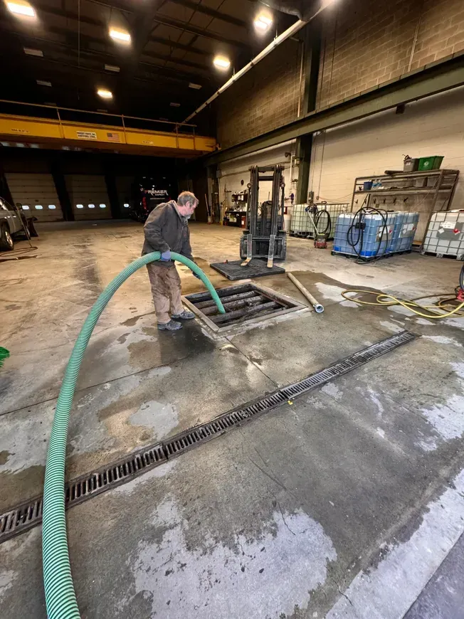 Man using a hose to clean a floor drain in a warehouse, near a forklift and equipment.