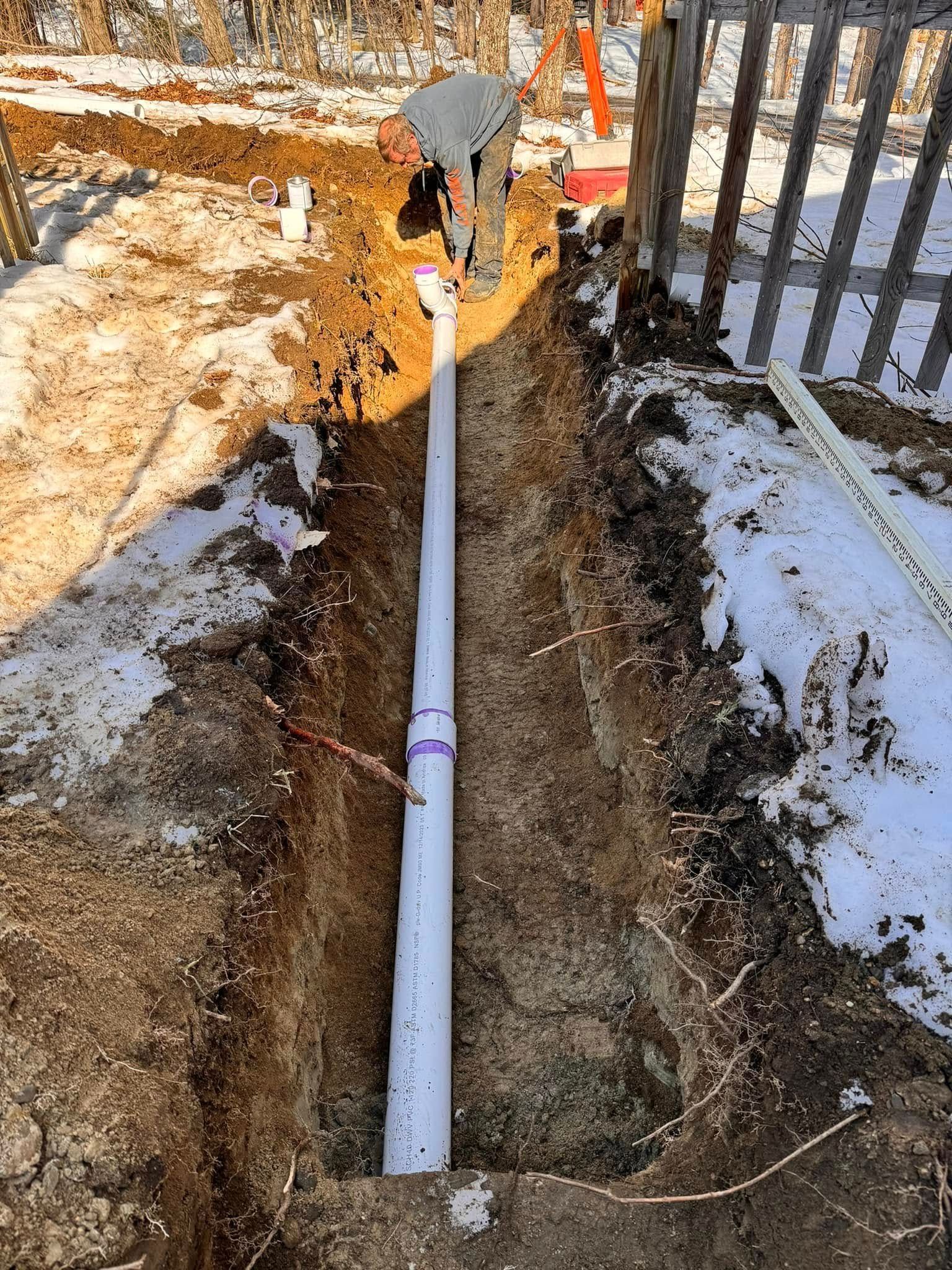 A trench with a white PVC pipe installed, near a wooden fence, in a snowy outdoor setting.