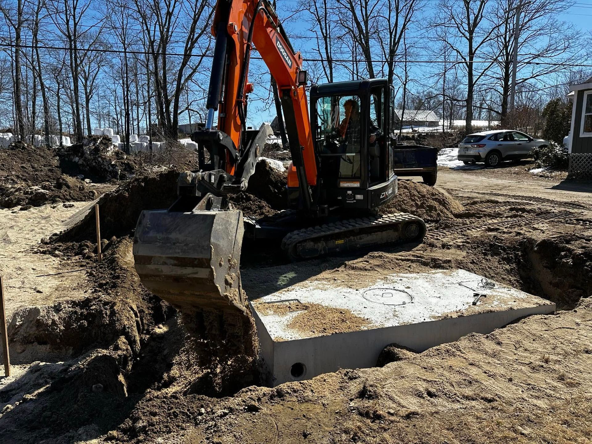 A small excavator digs around a concrete septic tank in a yard on a sunny day.