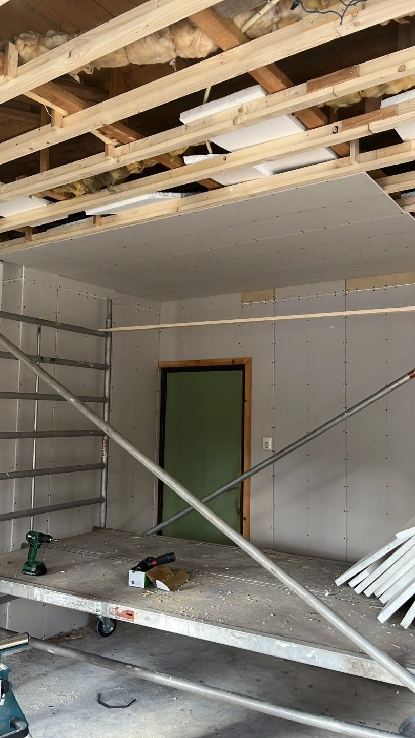 Gedeeltelijk gerenoveerde kamer met zichtbare plafondbalken, gipsplaten, steigers en een deuropening met groene kozijnen.