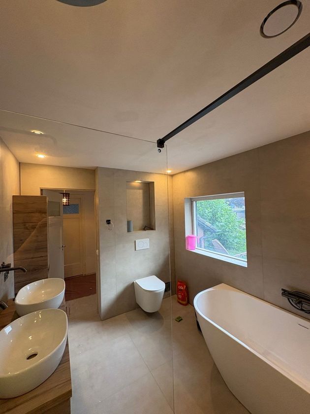 Moderne badkamer met dubbele wastafel, inloopdouche, vrijstaand bad en een klein raam met uitzicht op groen.