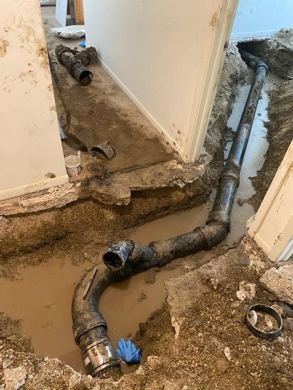 Pipes exposed in a trench dug into a floor, likely for plumbing repair, with mud and a blue glove.