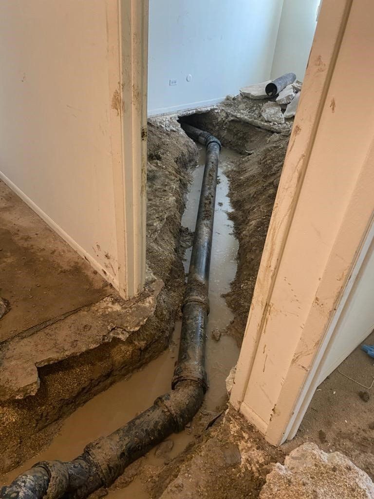 Trench dug through a room, exposing a long black pipe. The area is under construction; concrete and debris visible.
