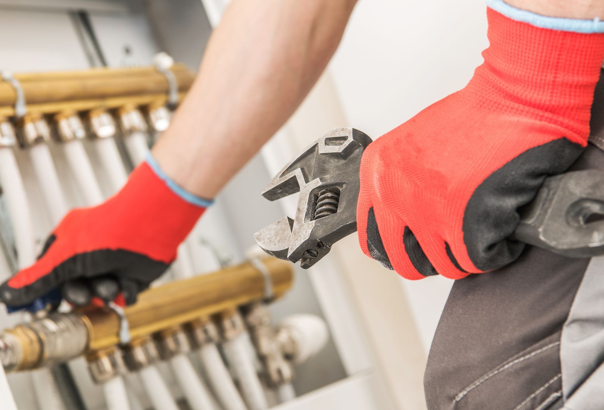 Photograph showing a close-up of a person wearing red gloves using an adjustable wrench to work on brass plumbing pipes and valves. Photograph showing a close-up of a person wearing red gloves using an adjustable wrench to work on brass plumbing pipes and valves.