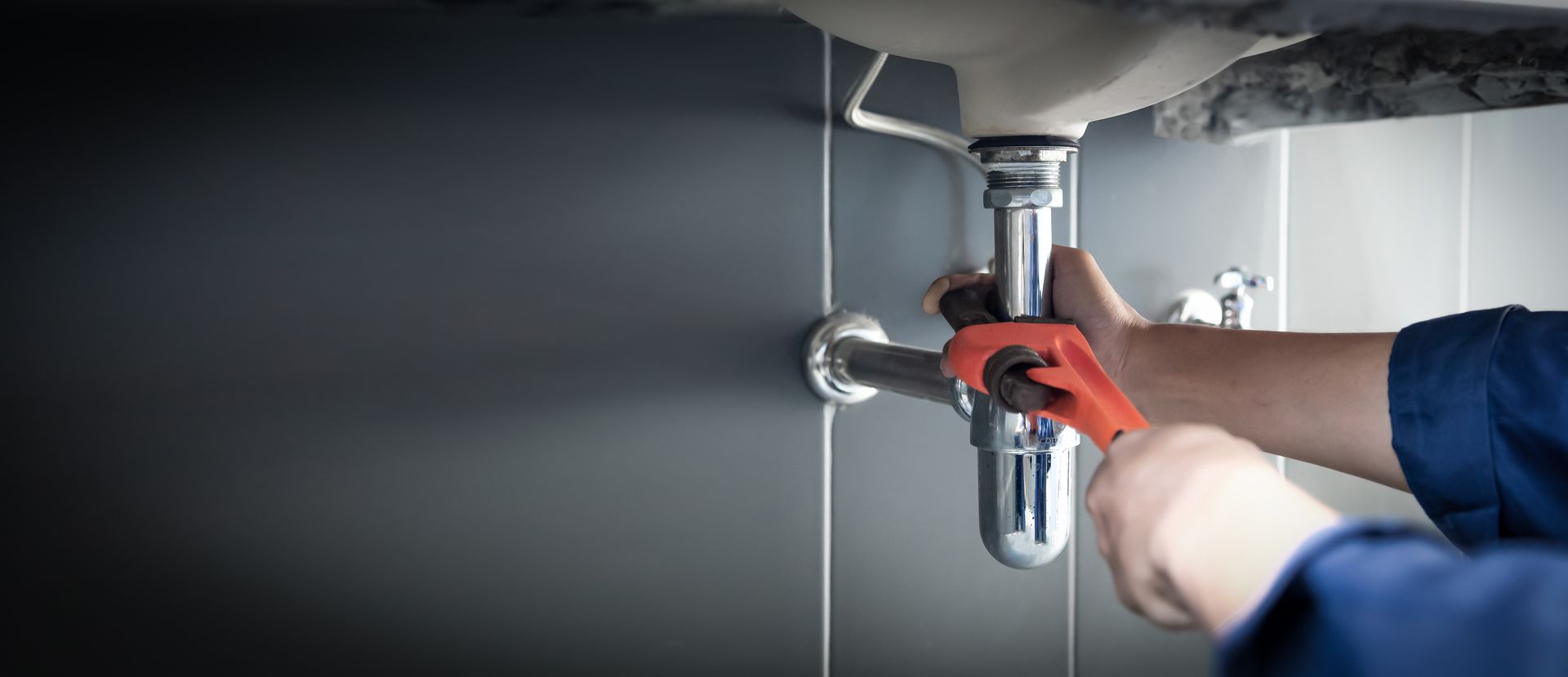 Plumber using a red wrench to tighten the chrome drain pipe under a white sink in a tiled bathroom.