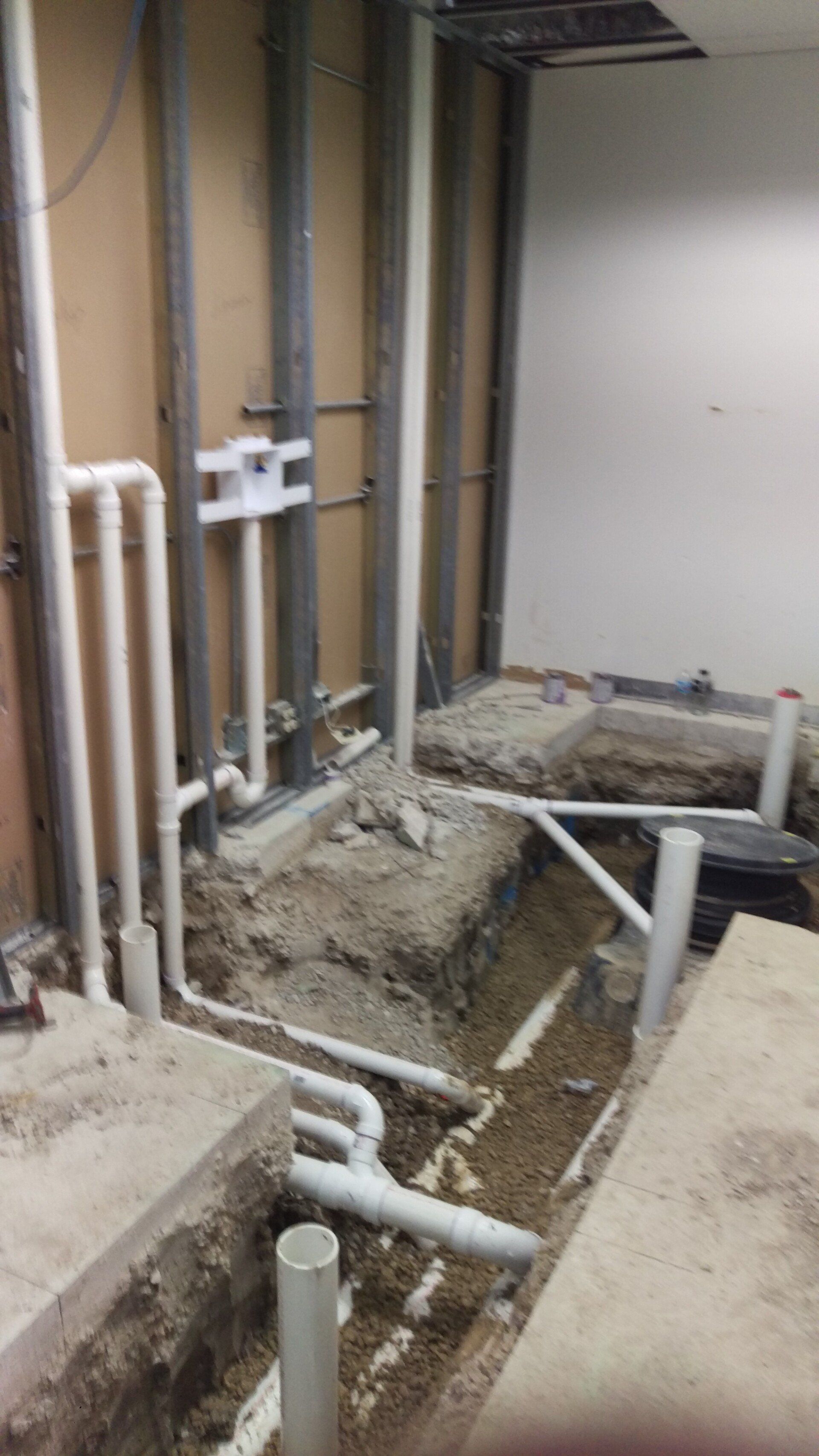 Plumbing work in progress: Excavated concrete floor, exposed pipes and metal studs in a room.