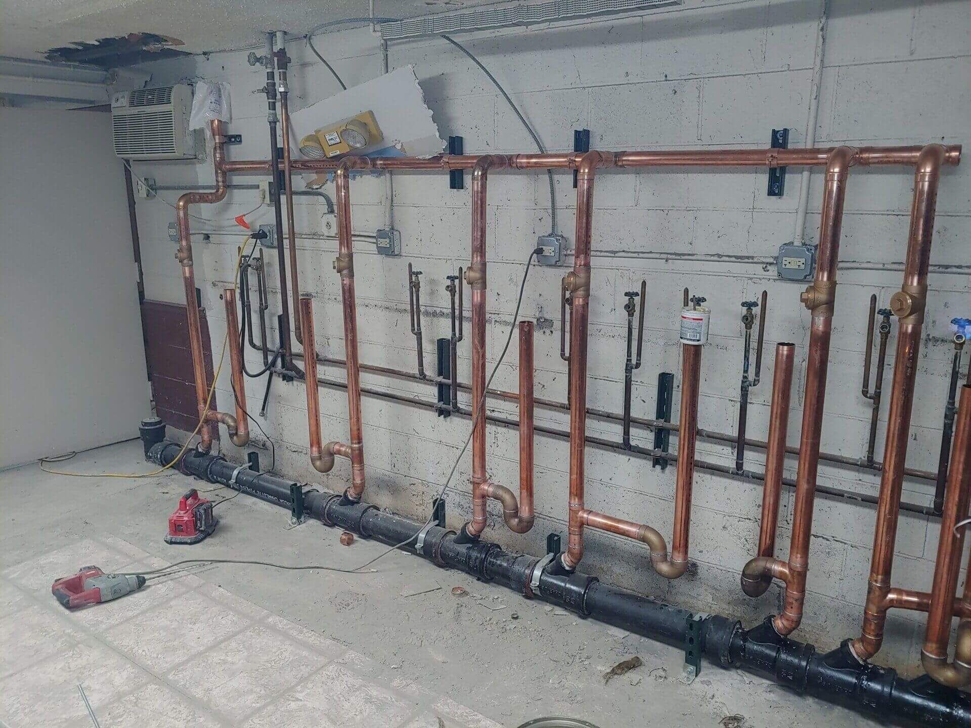 Copper pipes and black pipes installed on a white brick wall in a room under construction.