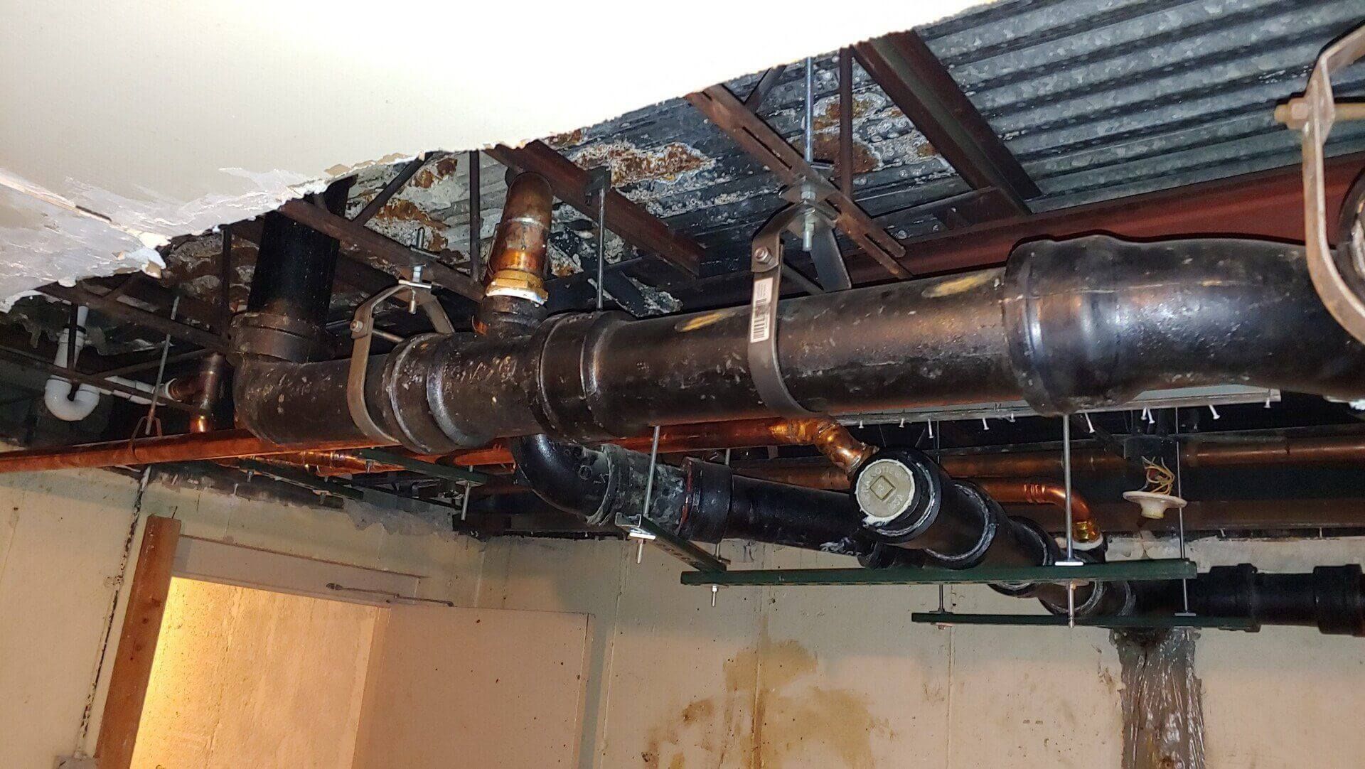 Pipes under a damaged ceiling. Black plumbing pipes with rust and water damage. Basement setting.