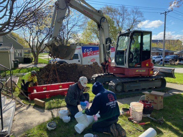 Construction workers replacing pipes on a residential lawn, using an excavator and tools. A white service van is in the background.