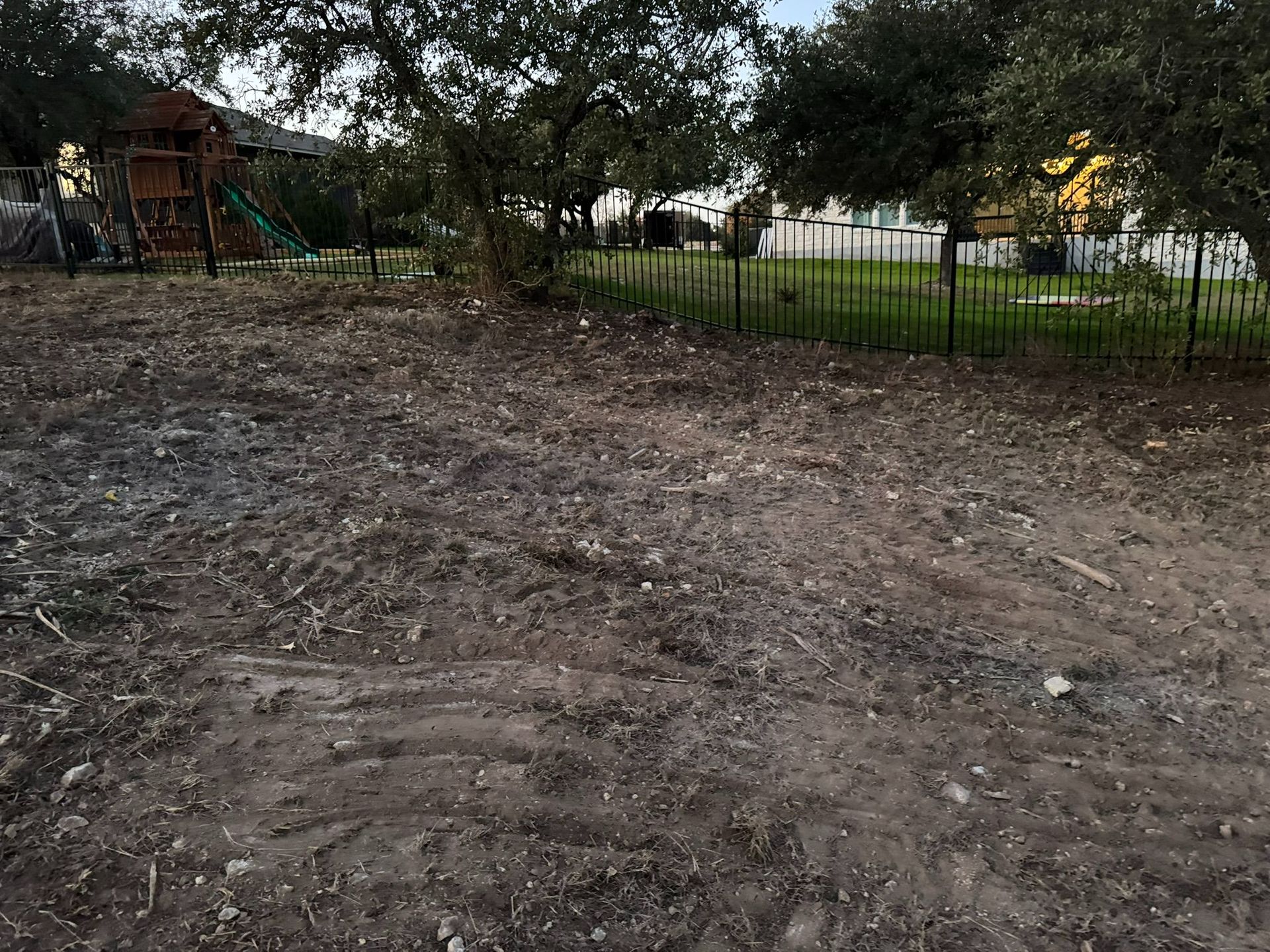 Dirt-covered yard with trees, a fence, and a playground structure in the background