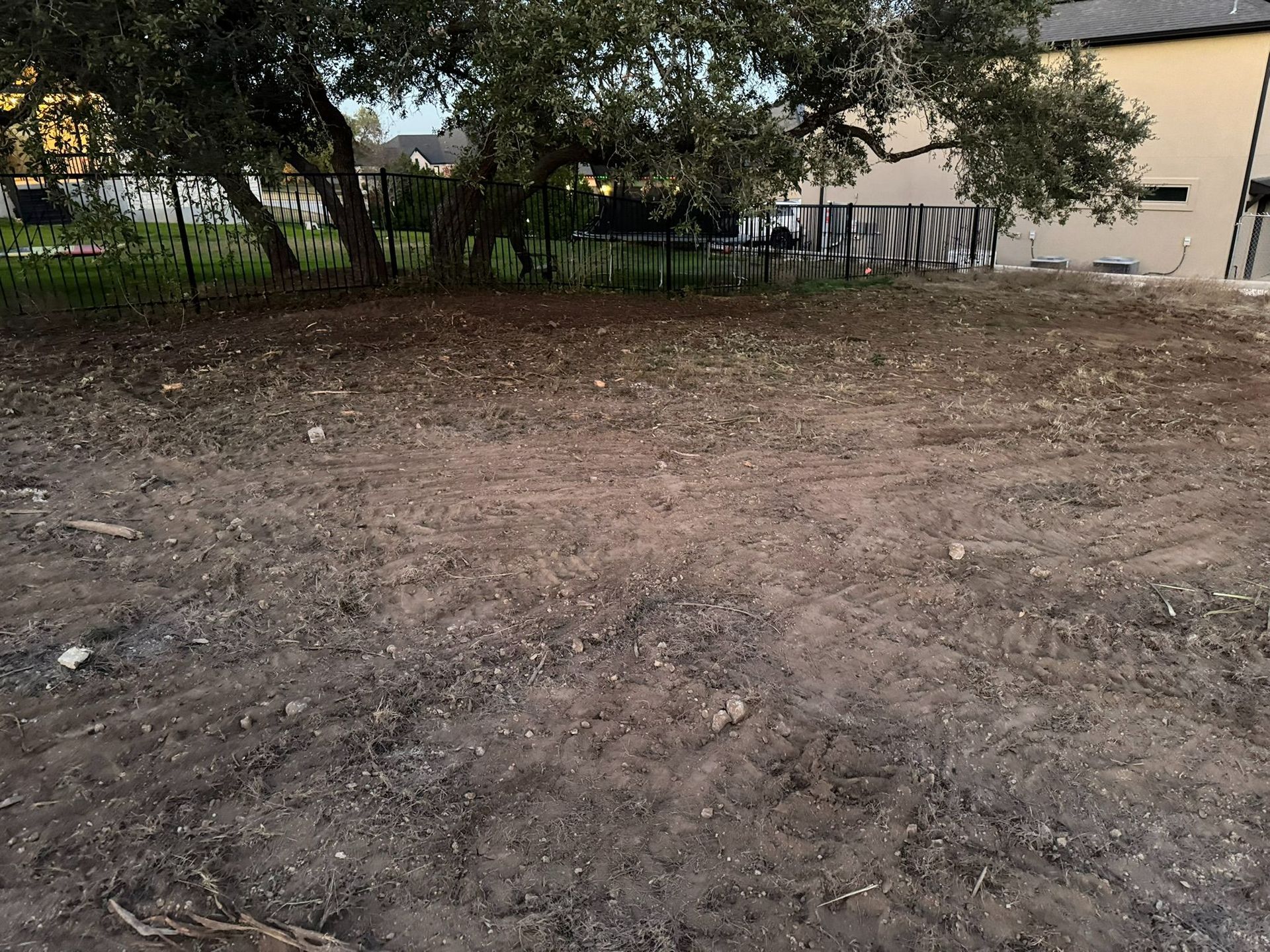 Empty dirt lot beside a beige building, with trees and a fence in the background.