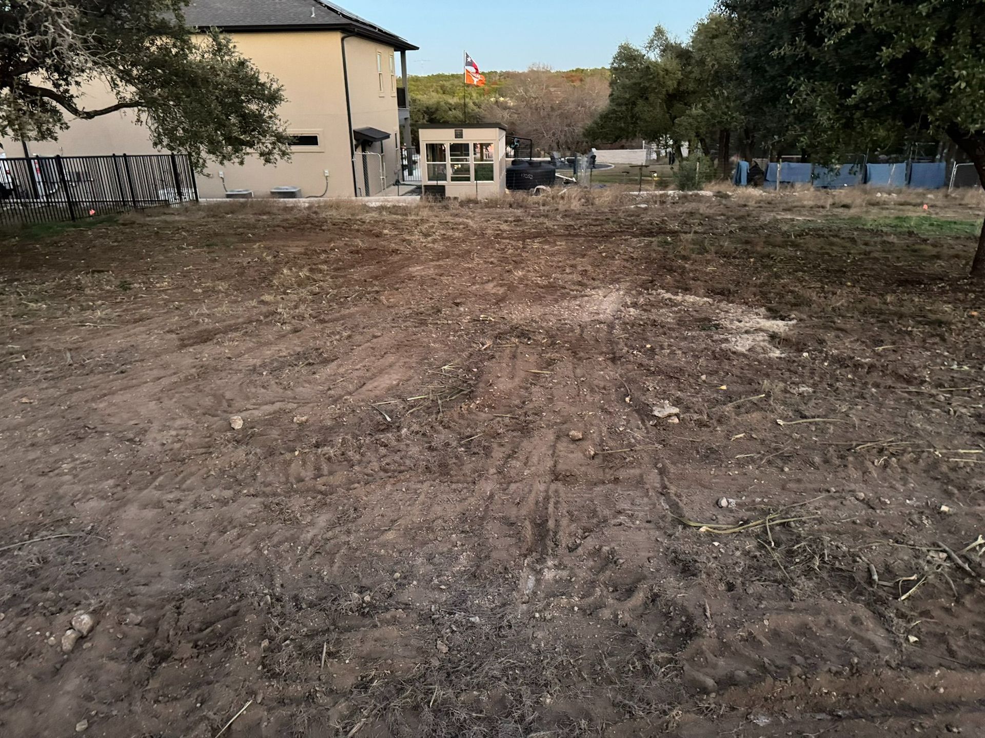 Dirt lot beside a beige building, with trees and a small structure in the background.