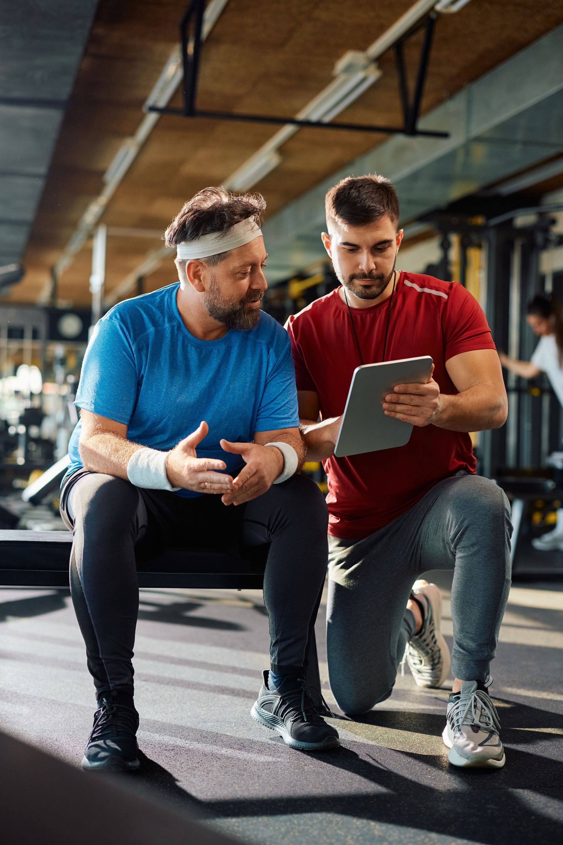 A man and trainer in a gym discuss progress using a tablet. Man wears blue; trainer wears red.