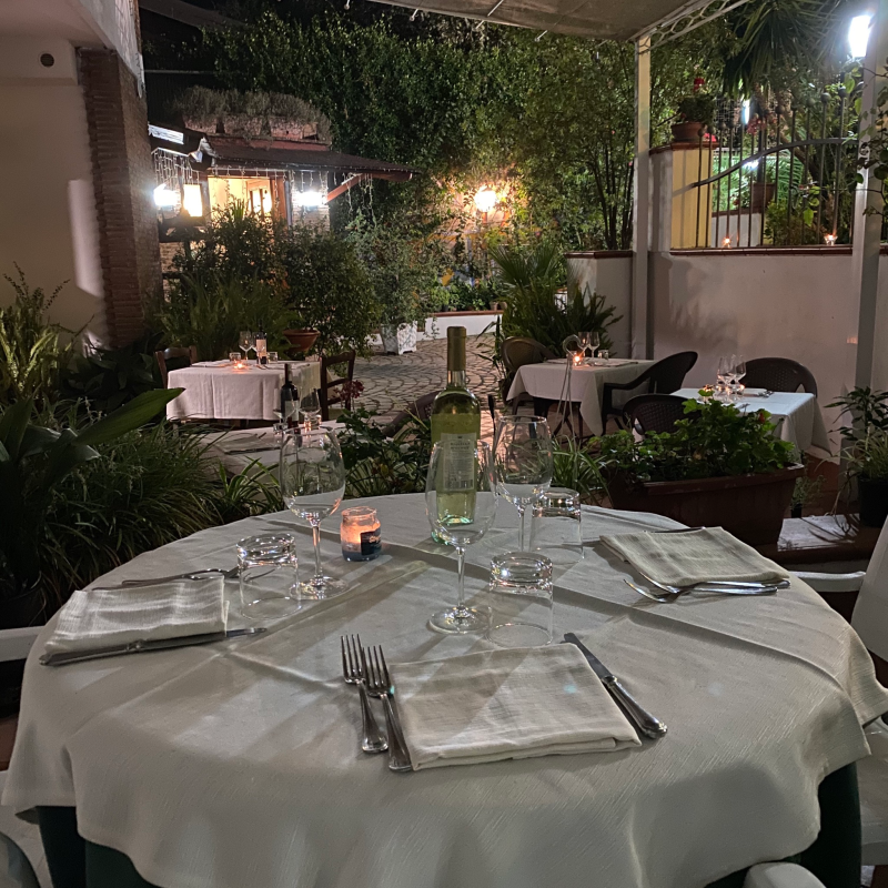Ristorante all'aperto con tavoli apparecchiati per cenare. Tovaglie bianche, candele, verde, allestimento serale.
