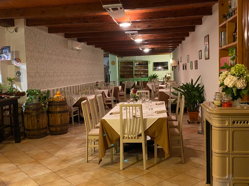 Interno del ristorante con tavoli apparecchiati per la cena. Travi in legno, pareti bianche e pavimento piastrellato.